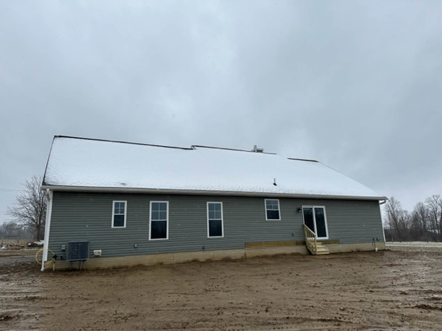 Two-story house with white snow covering the roof, white-framed windows, gray siding, and bare trees in the yard