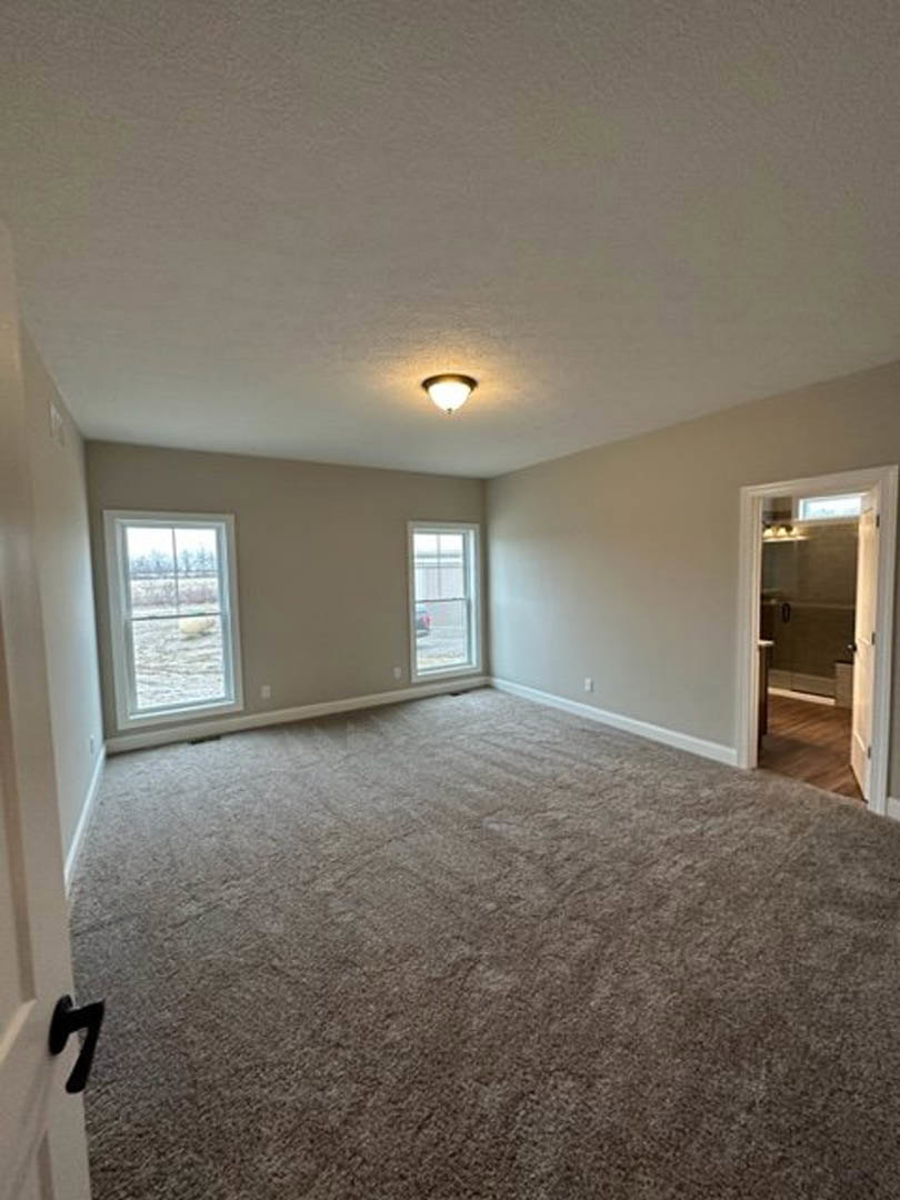 Bedroom with brown carpet, open door, two windows showing snowy field and parked car, white walls, ceiling-mounted light fixture