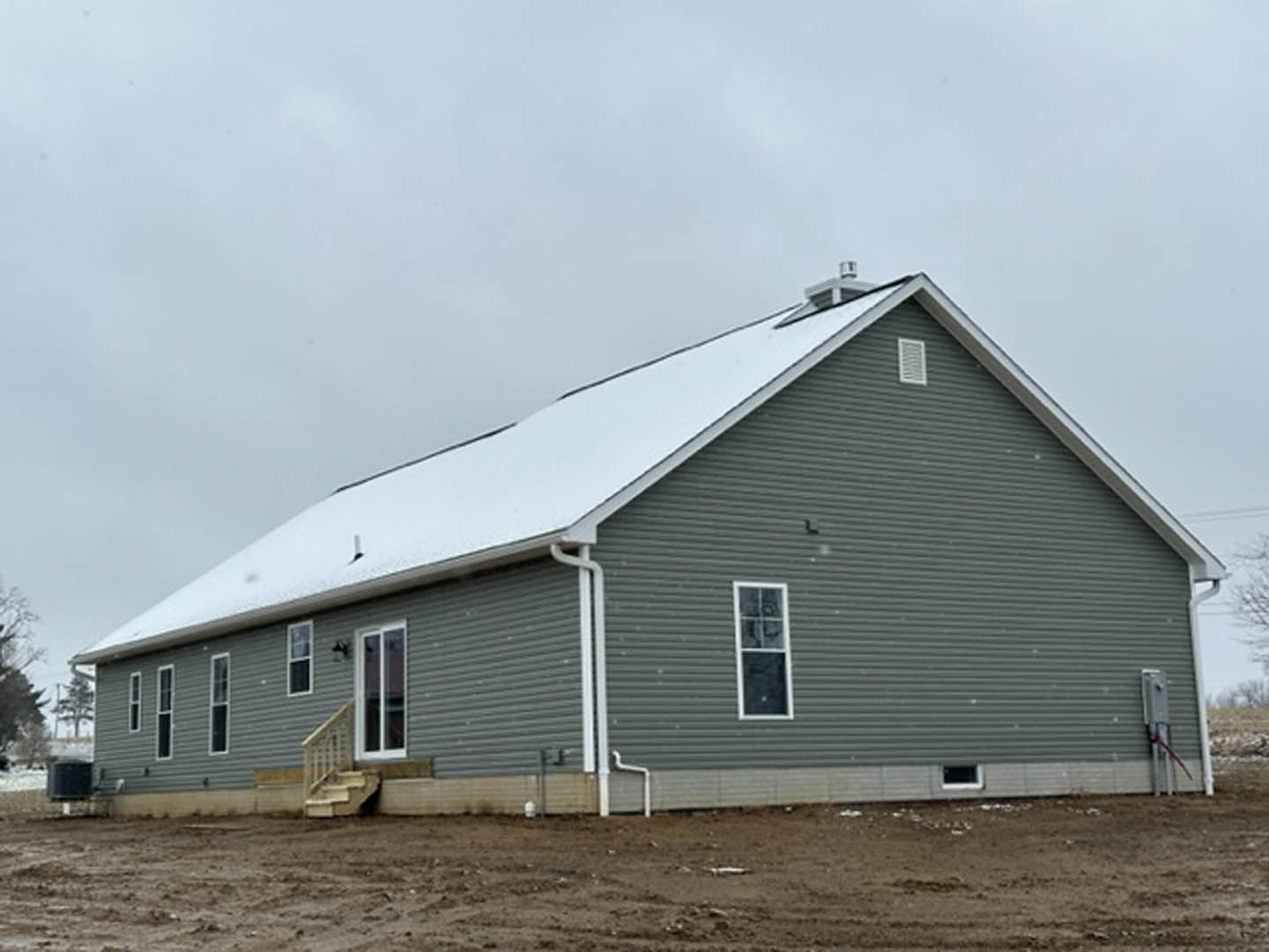 Two-story cottage with white siding and snow-covered roof, surrounded by a dirt field and wooden fence under a cloudy sky