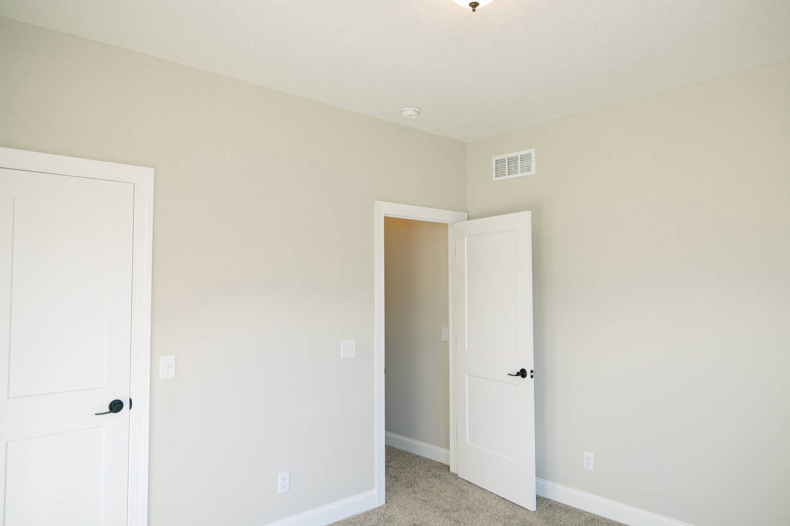 White paneled door with matte black handle open to reveal light gray walls, white baseboards, and vent; hardwood flooring and wall-mounted light switch visible