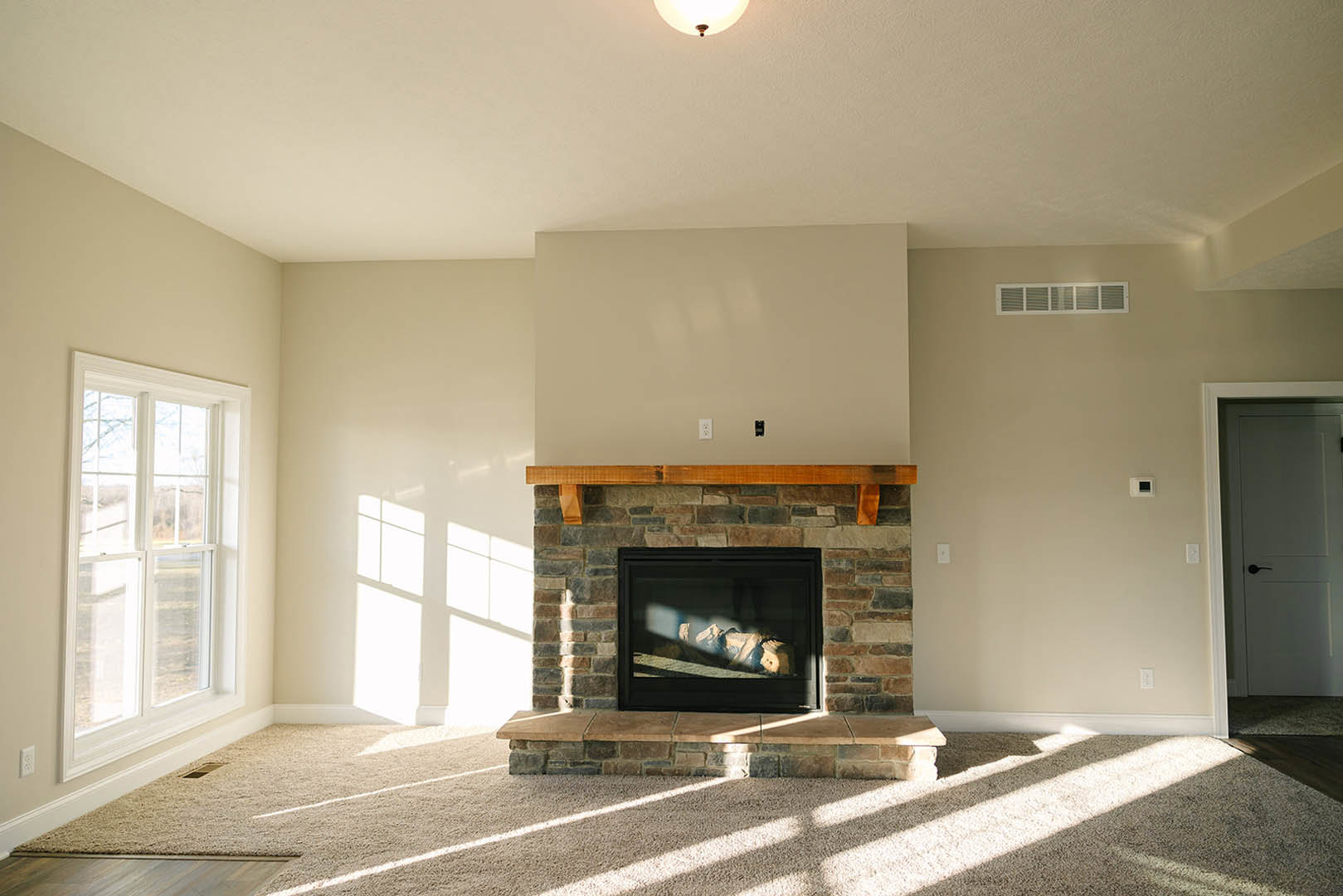 Stone fireplace with glass door and stacked wood, plaster walls, hardwood flooring, large window offering outdoor view, partial door and stone step visible
