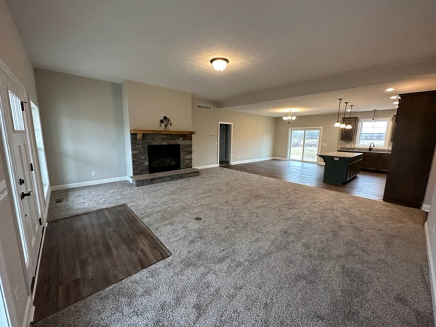 Open-concept living room with wood flooring, stone fireplace, recessed lighting, and adjacent modern kitchen with white cabinetry and stainless steel appliances