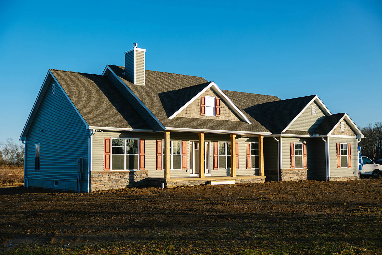 Two-story farmhouse with white siding, large covered porch, expansive grassy yard, white-framed windows, and a white van parked on a dirt driveway.