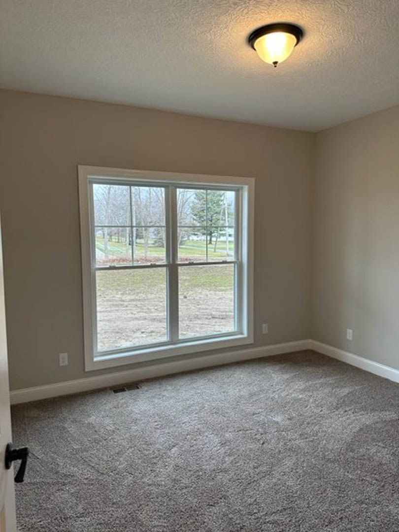 Neutral-toned carpeted room with white walls, large window overlooking grassy field, ceiling-mounted light fixture, and brushed metal door handle