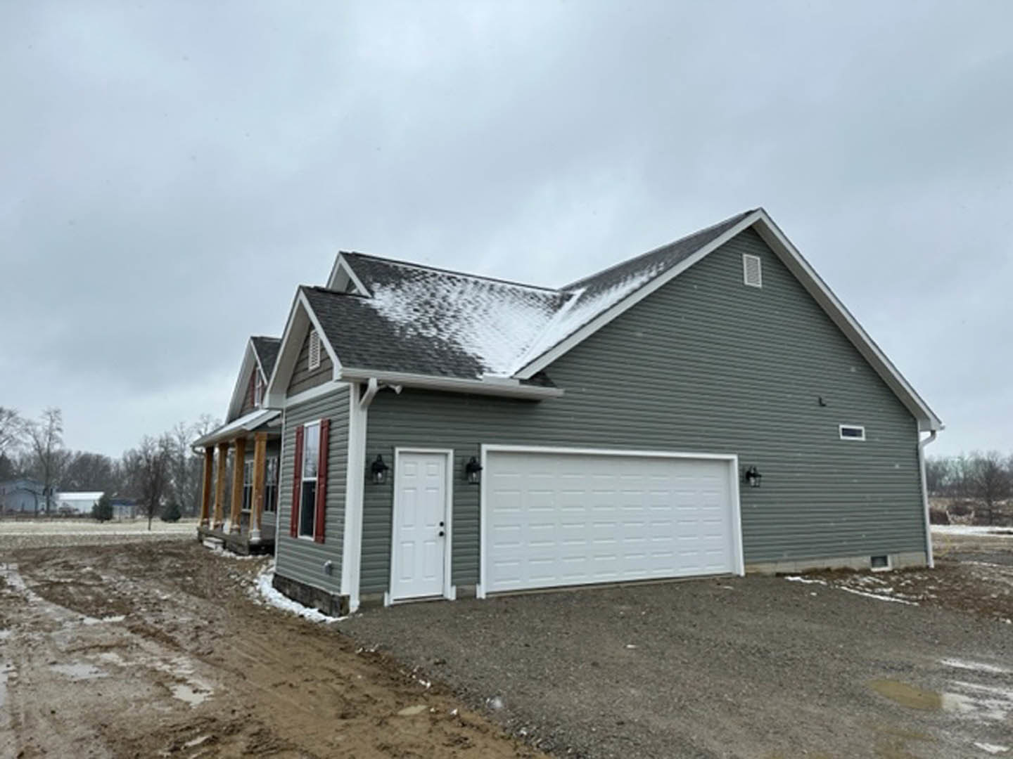Two-story house with attached garage, white siding, black hardware, snow-covered roof, dirt driveway, and overcast sky