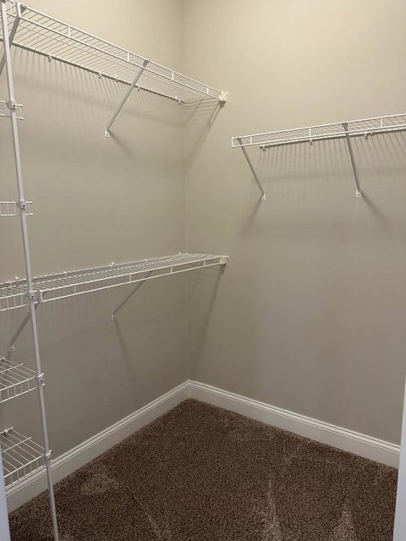 Walk-in closet with white metal shelving mounted on walls, beige carpet flooring, and neutral painted walls