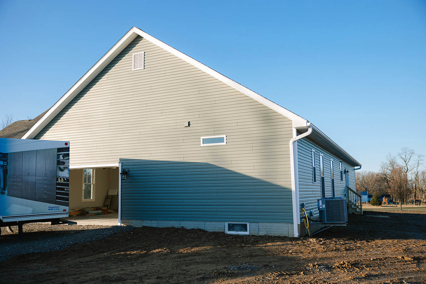 Two-story house with attached garage, blue siding, white trim, large windows, and a paved driveway; mature trees and lawn in the foreground.