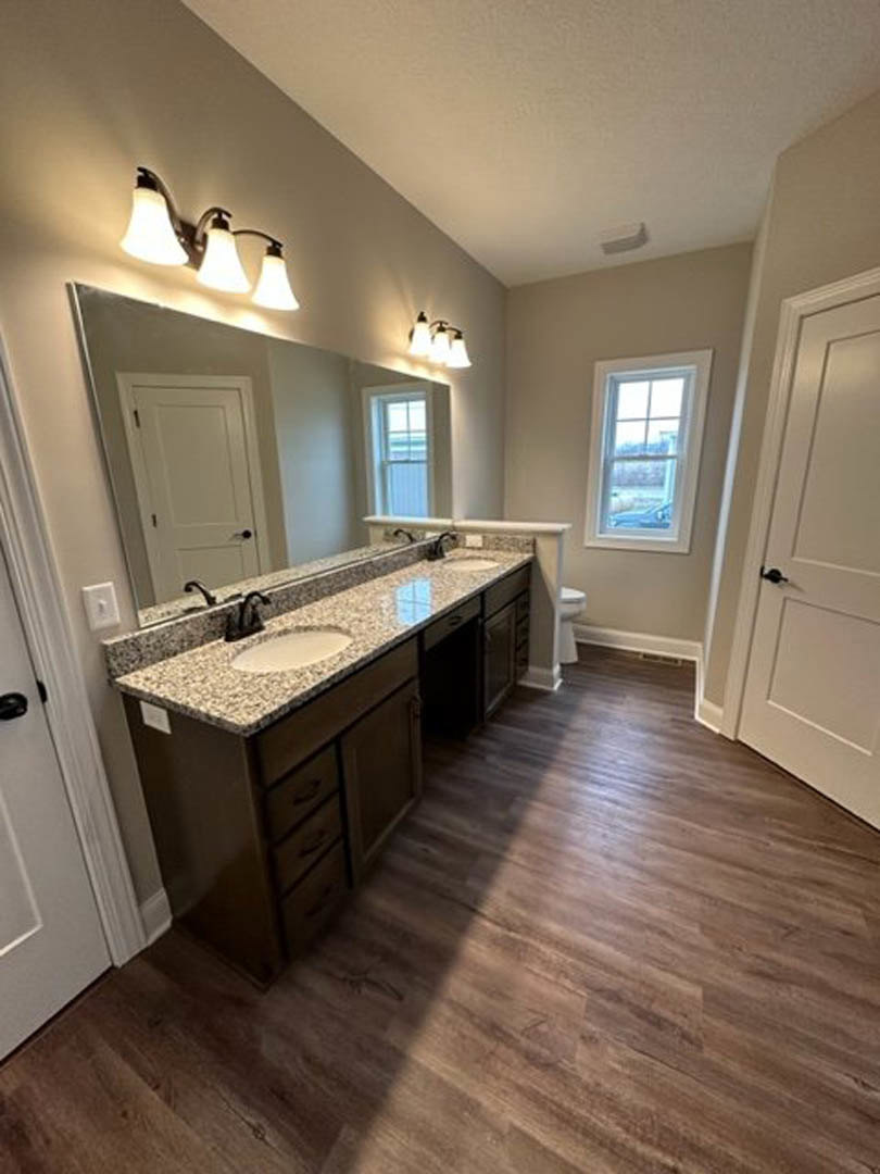 Bathroom with double vanity, wide mirror above, light fixture with three bulbs, wood flooring, tile walls, cabinetry, and window overlooking cityscape.