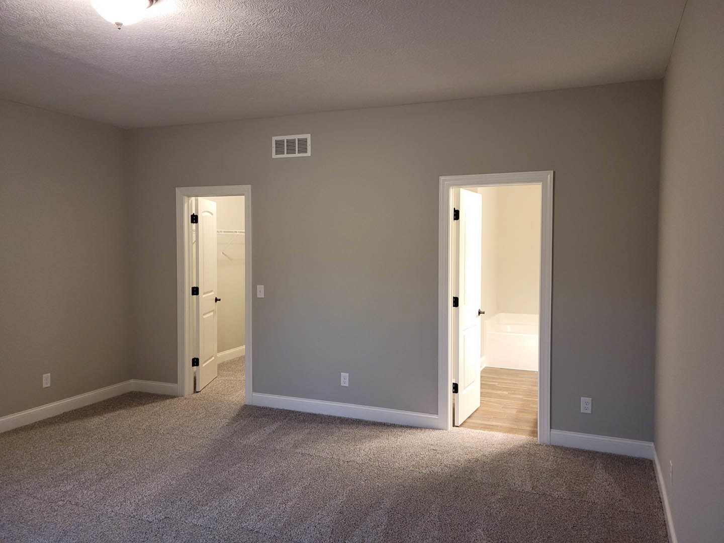 Bathroom with freestanding white bathtub, two white paneled doors with black hardware, carpeted floor, wood flooring, wall vent, and overhead lighting.