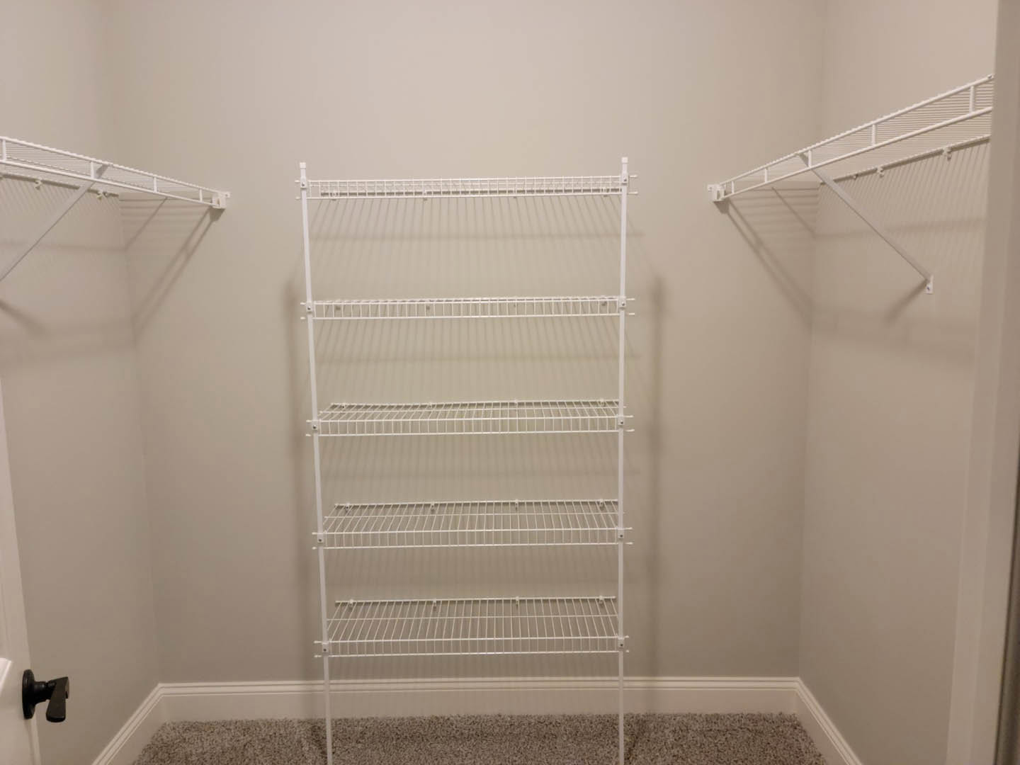 White wire shelf mounted on a white wall above beige carpet, black door handle visible on adjacent wall.