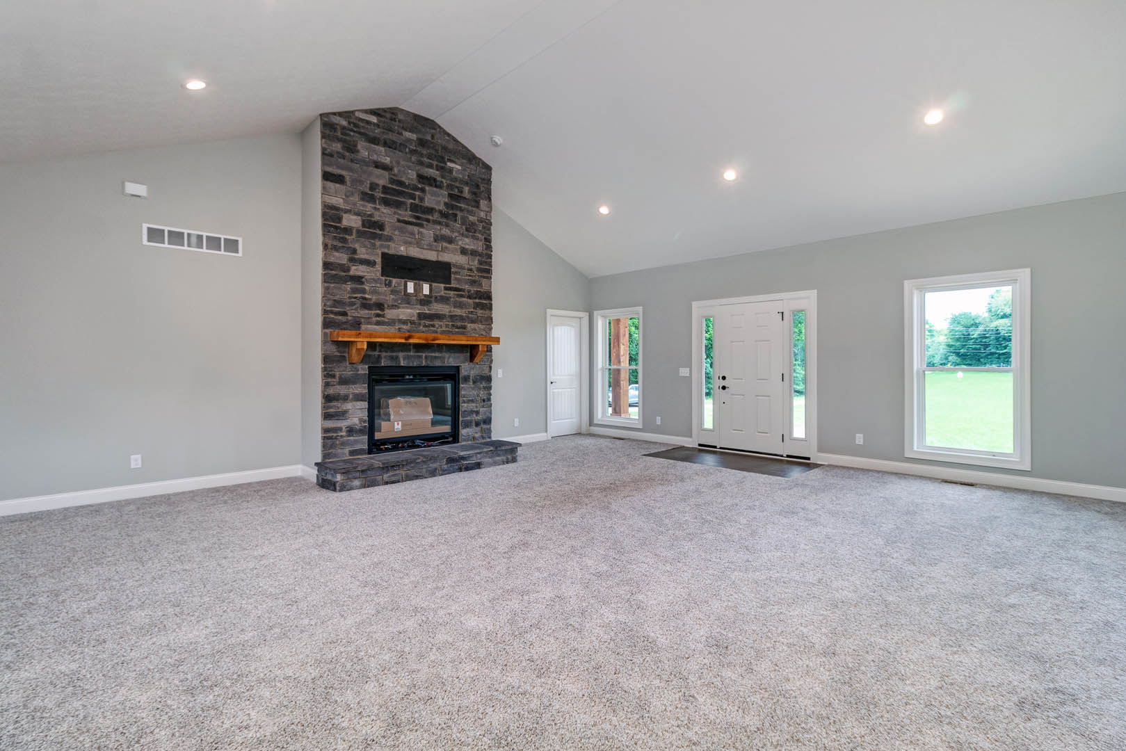 Living room with beige carpet, brick fireplace featuring a wooden mantel and a box inside, large window overlooking green lawn, white door with glass panes, light-colored walls and