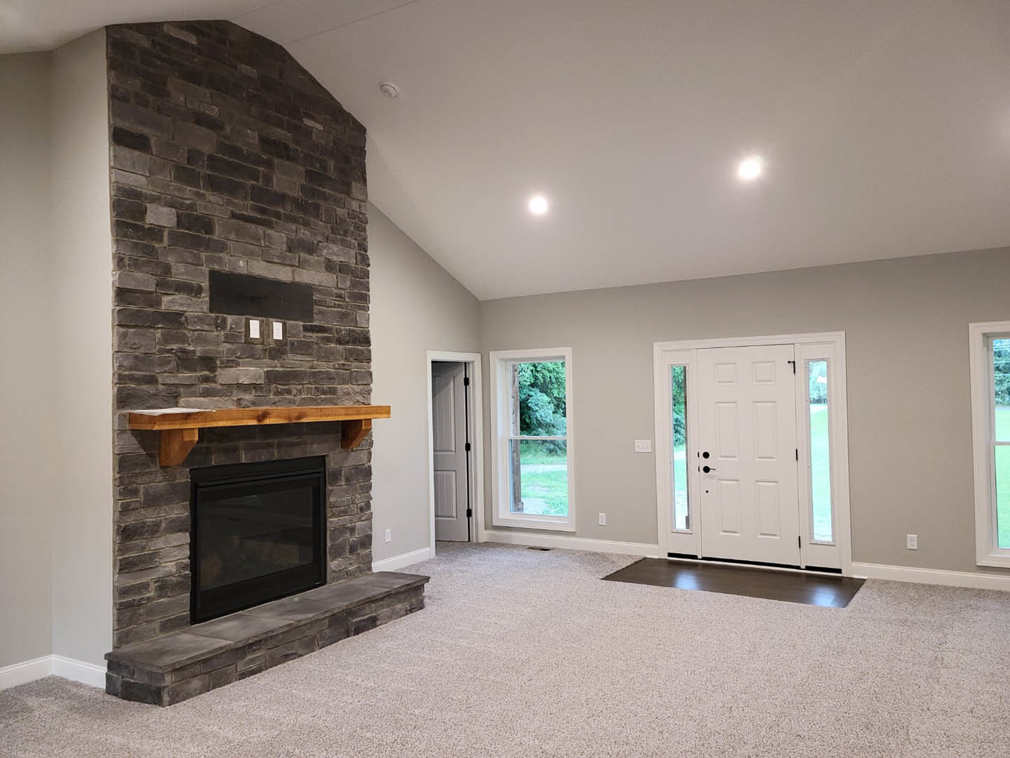 Black wood-burning fireplace with glass door set in a brick wall, wood mantel shelf above, white door with glass windows, large window showing trees outside, hardwood flooring and