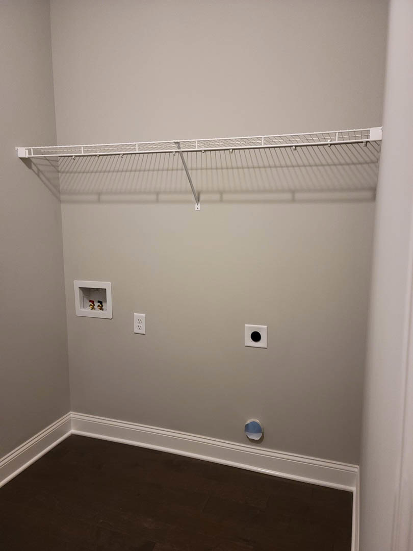 White built-in shelf against plaster wall, dark wood flooring with white baseboard, metal rod and white utility box with valves, ceiling visible above.