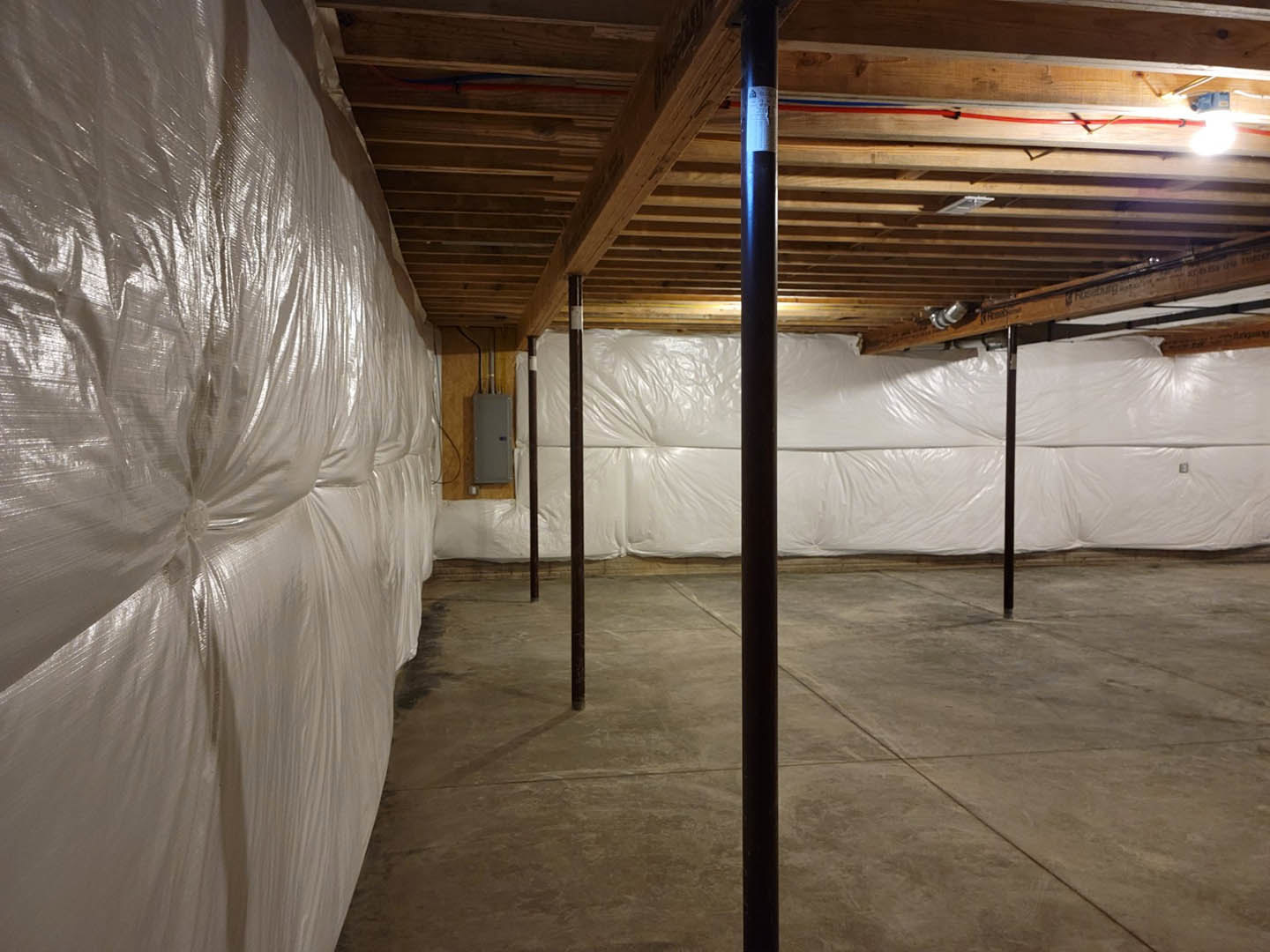 Basement with wooden ceiling, white plastic sheeting covering walls and bed, concrete floor with metal pole, white box featuring black buttons
