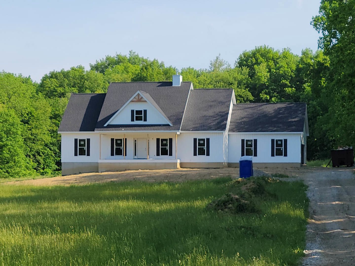 Two-story house with black roof, white siding, and square windows, set on a large grassy lawn with mature trees and historic site visible in the background; stone path leads across