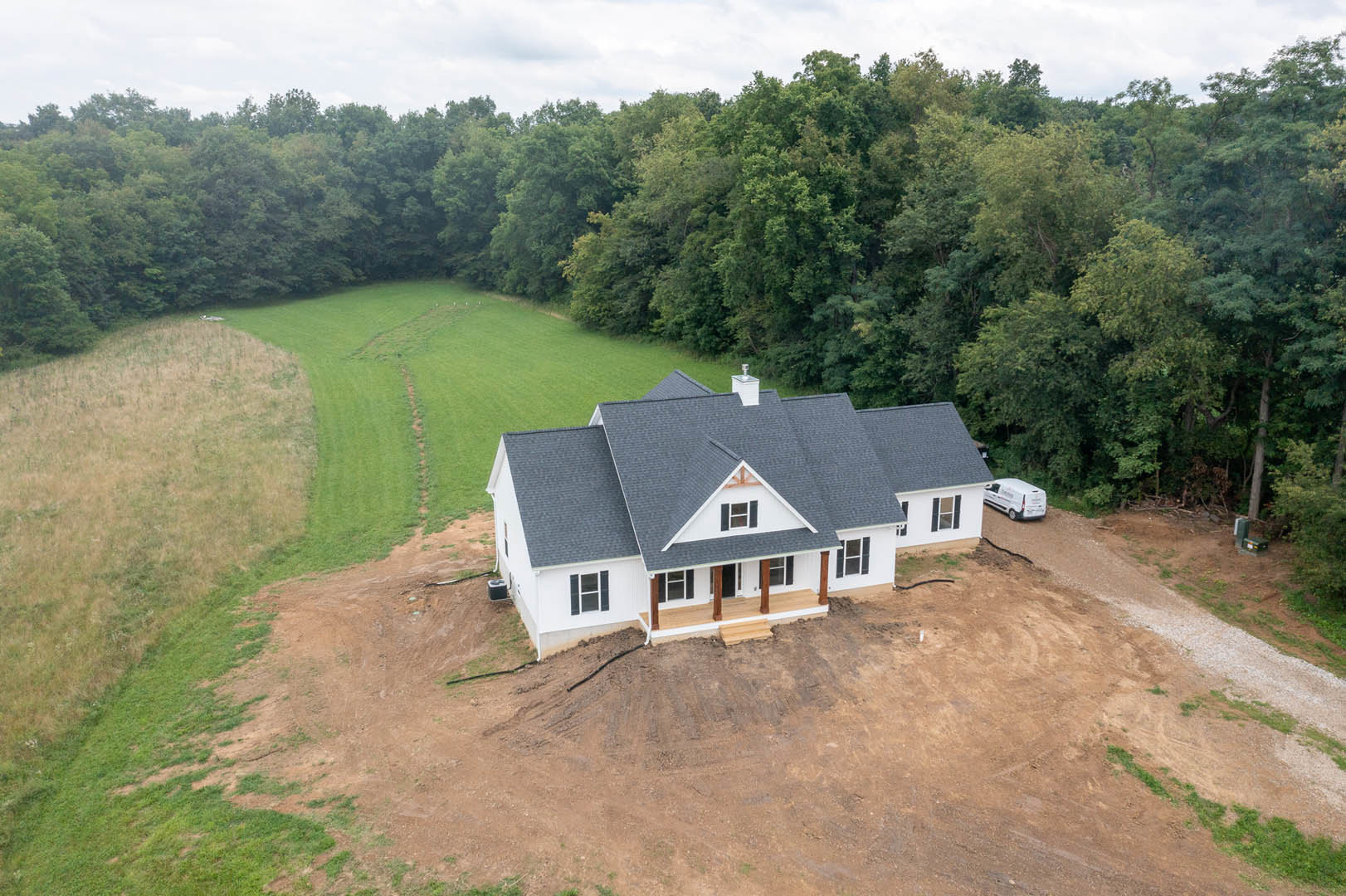 Two-story house with unfinished exterior, large covered porch, surrounded by grassy field and scattered trees, white van parked near dirt driveway, cloudy sky overhead