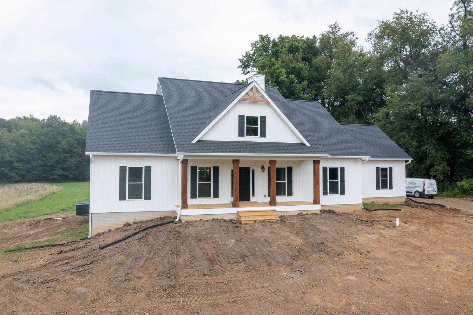 Partially built house with black roof, exposed framing, muddy ground, surrounding trees, white van parked nearby, window with shutters visible on side
