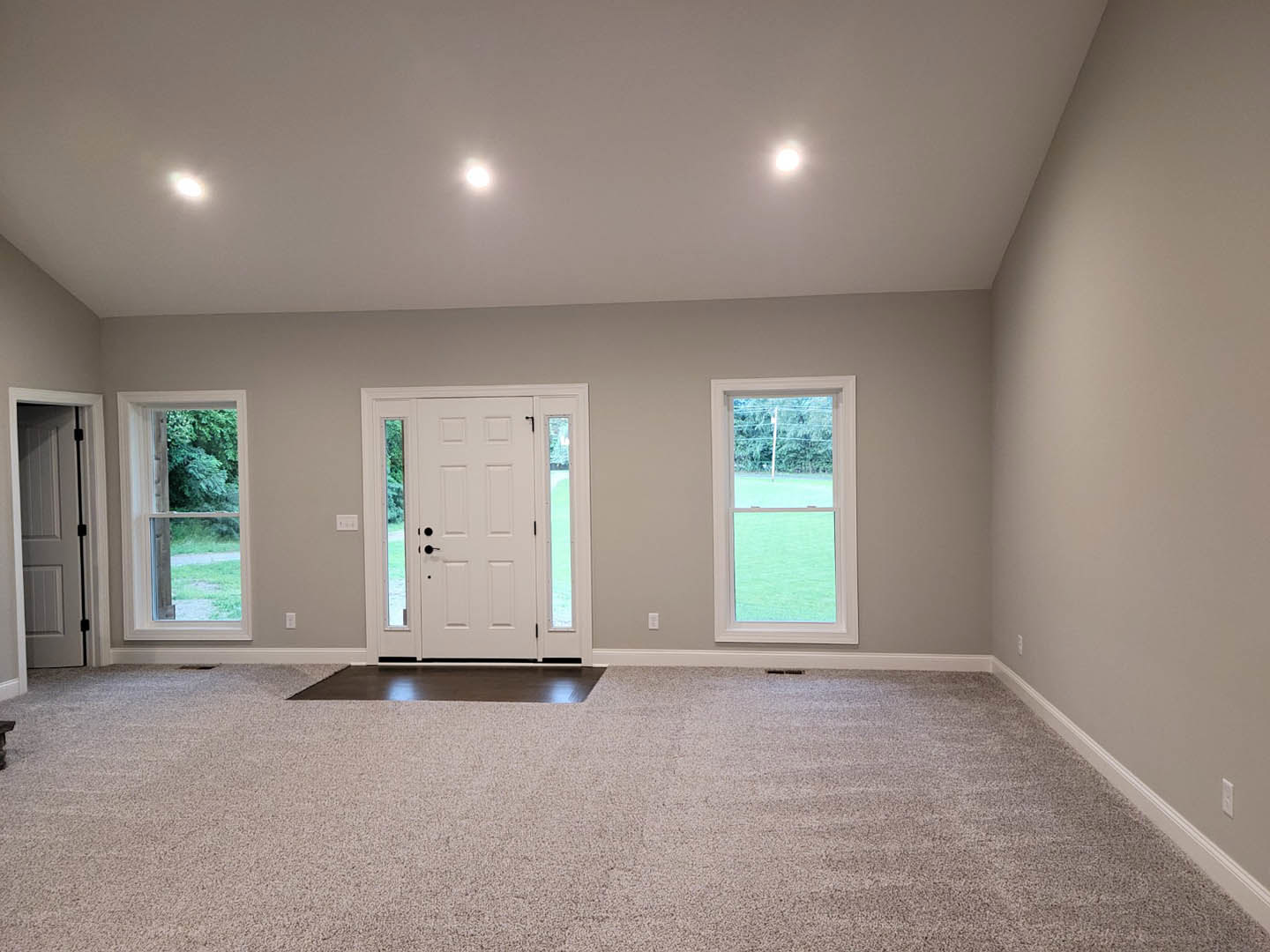 Carpeted room with white walls, white door featuring glass panels and black handle, large window overlooking green lawn, ceiling with recessed lighting