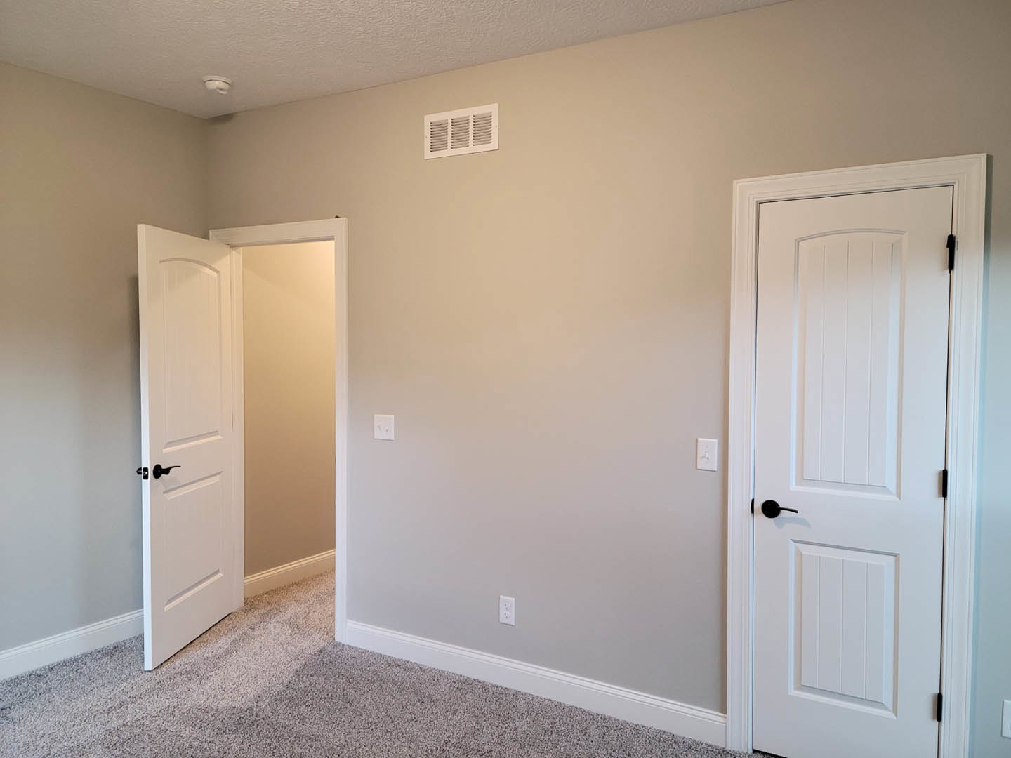 White interior room with two doors featuring black handles, light switches on the wall, wall vent, and neutral flooring.