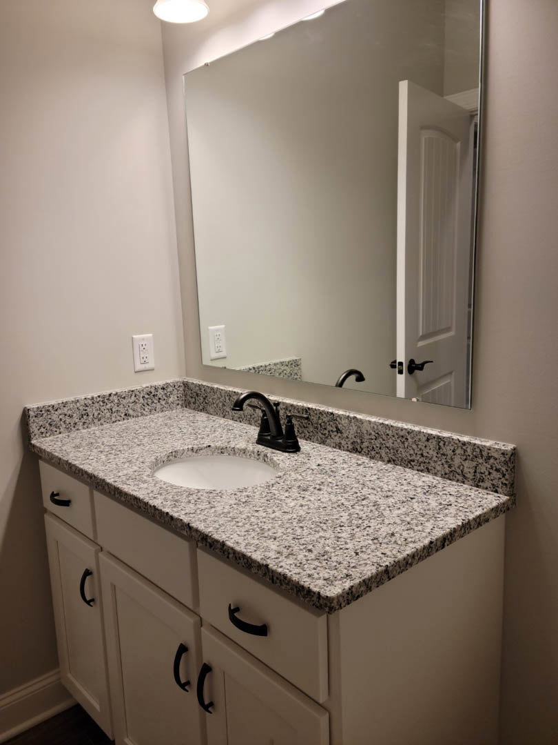 White ceramic sink with matte black faucet set on a light stone countertop, rectangular wall mirror above, gray tile backsplash, and wood cabinetry below.