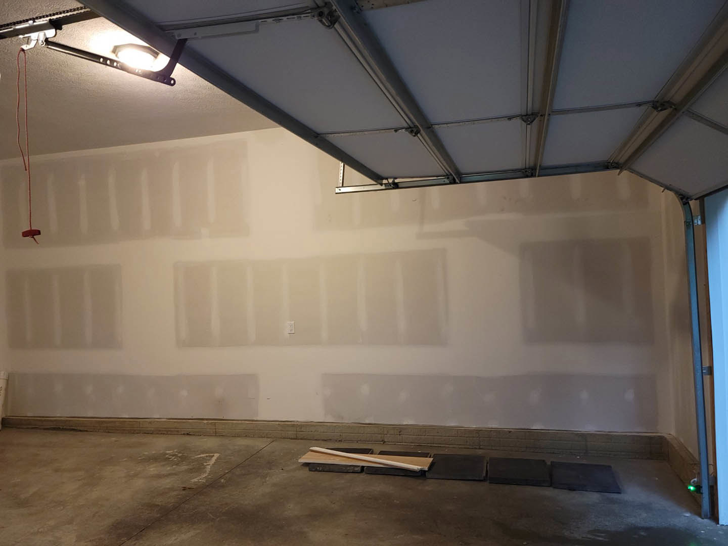 Garage interior with smooth white plaster walls, white ceiling panels, overhead light fixture, wood plank detail, and red wire on the floor.