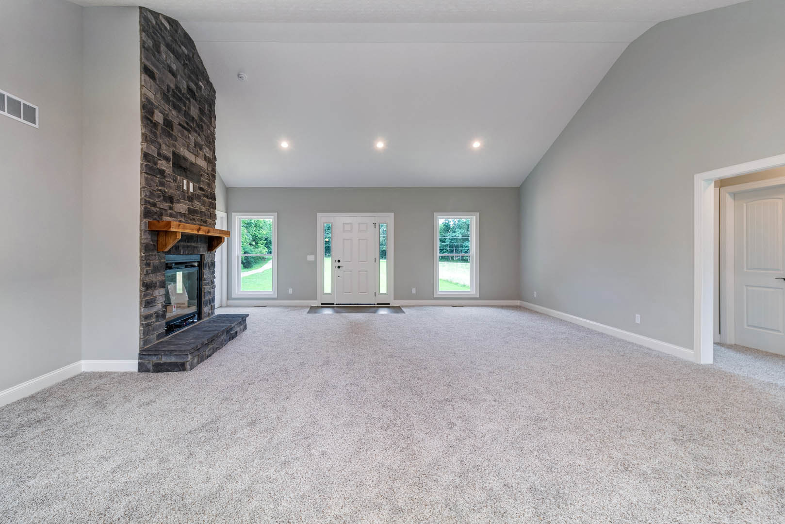 Living room with beige carpet, stone fireplace, white door with glass panes, large windows showing trees outside, plaster walls, and crown molding.