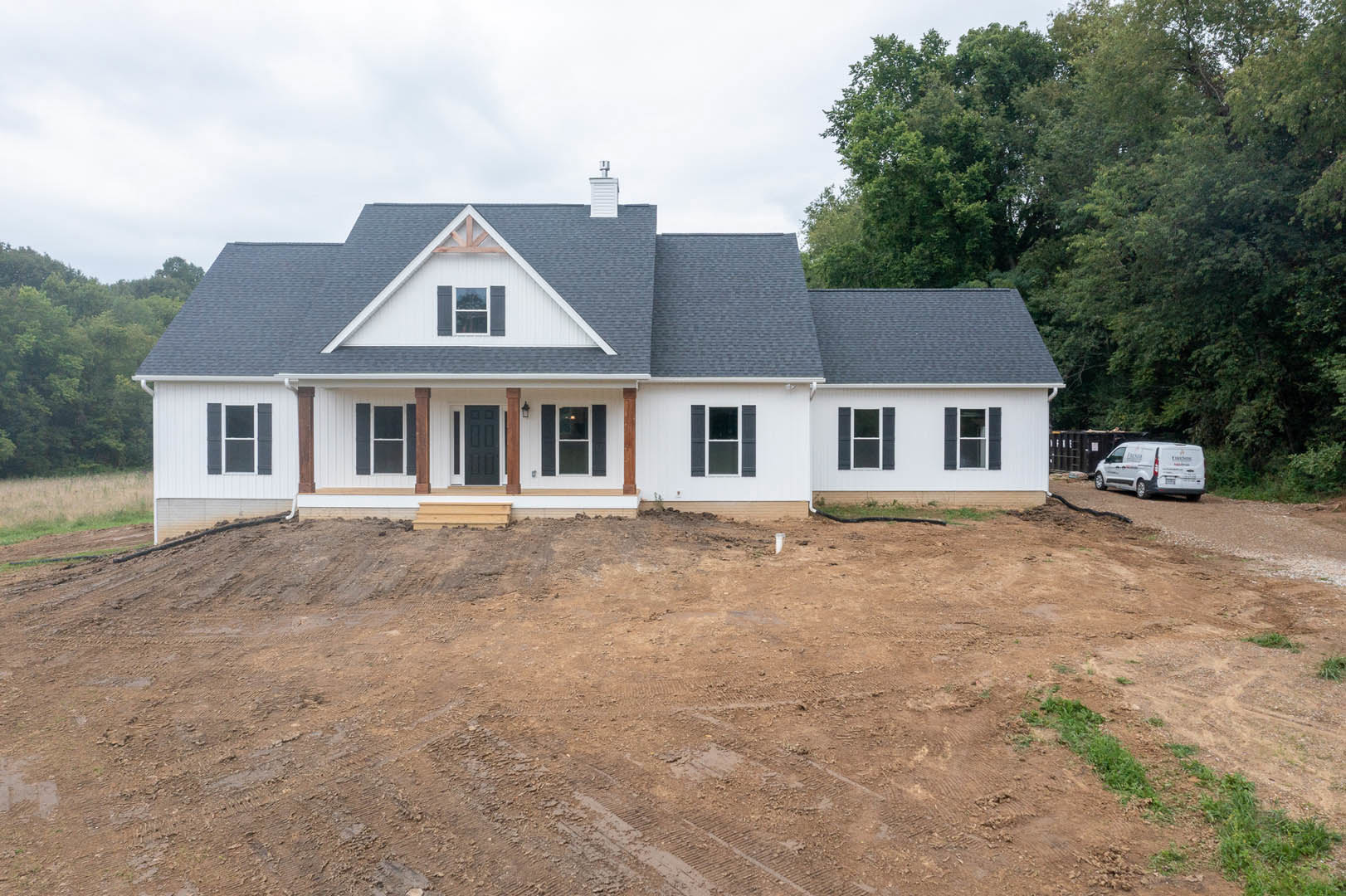 Two-story house under construction with exposed dirt yard, surrounding trees, black front door with white frame, white-framed windows, and brick chimney