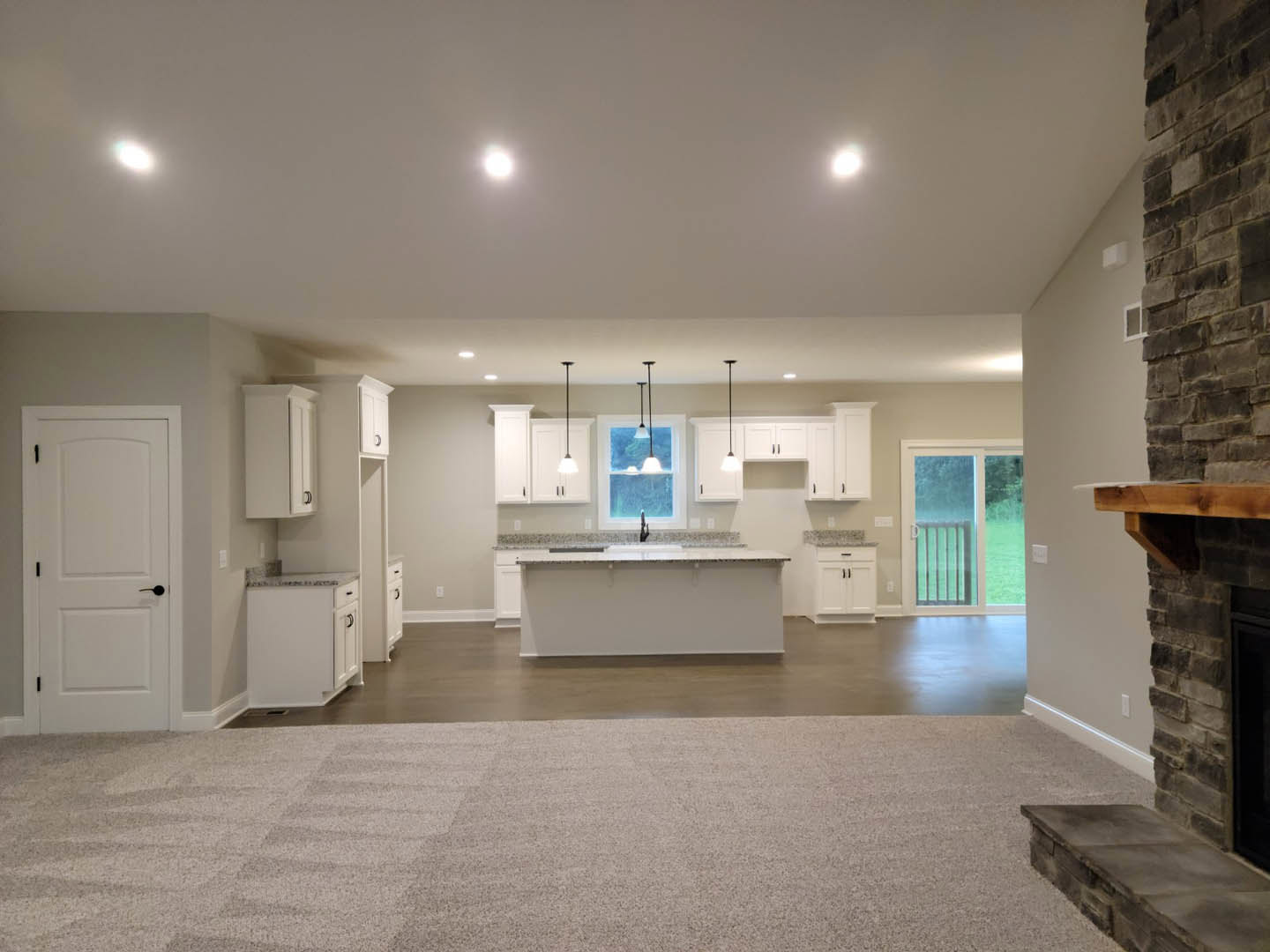 Open-concept kitchen and living room featuring a stone fireplace, white cabinetry, black hardware, white walls with floating shelves, sliding glass door leading to fenced yard