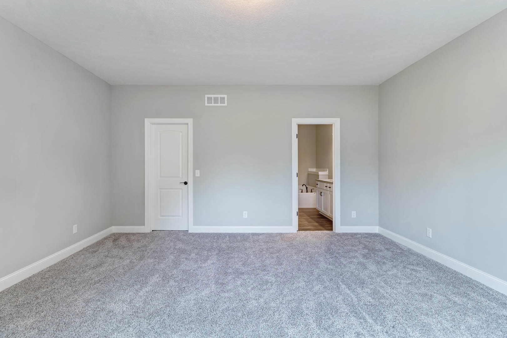 Bedroom with light gray carpet, white paneled door featuring a black handle, adjacent bathroom with white vanity, sink, and bathtub, soft overhead lighting highlighting smooth