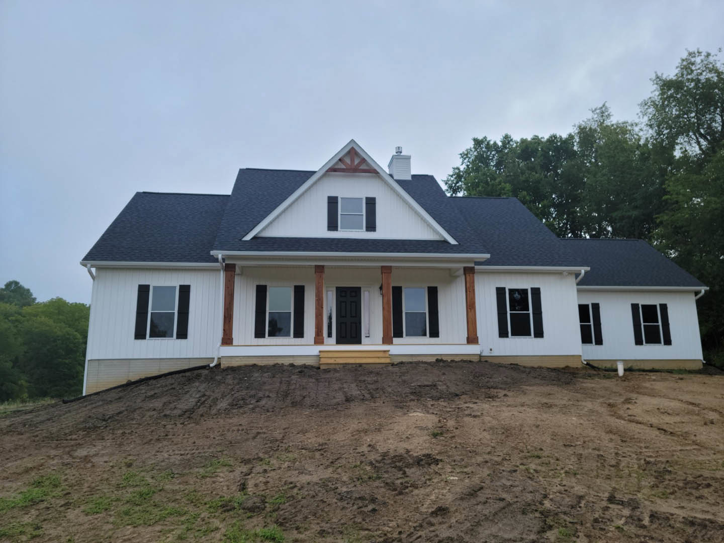 White house with black roof and black door, black window shutters, wooden step leading to dirt field, hill and trees in the background