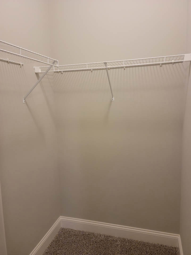 White built-in shelf with metal hanging rod mounted on a plaster wall in a closet, beige carpet flooring visible.