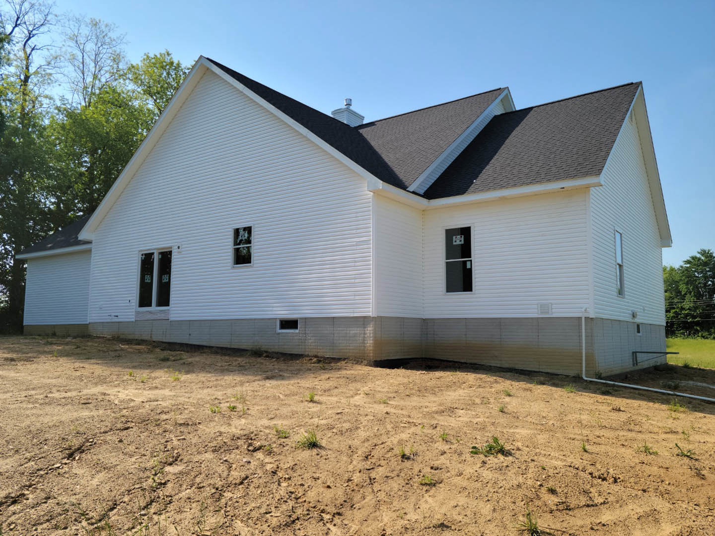 White house with black roof, white-framed windows, surrounded by dirt and sparse grass, trees in the background, clear blue sky overhead