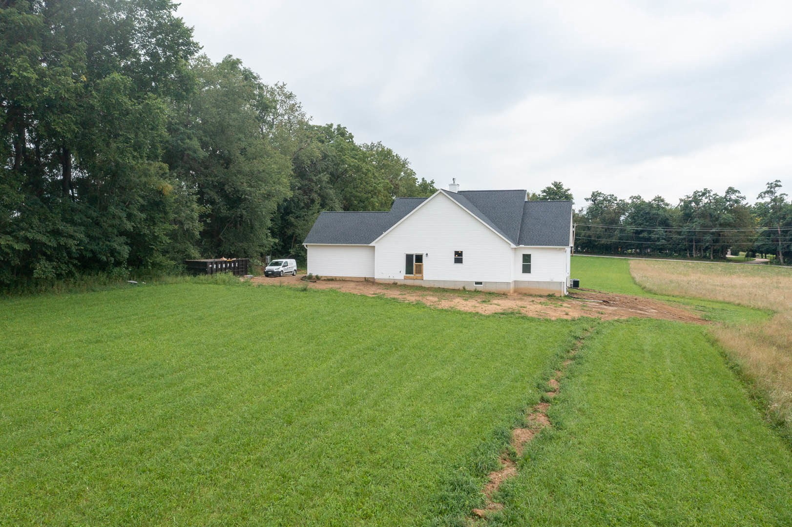 White house with black roof set on expansive green lawn, bordered by trees and open field under partly cloudy sky