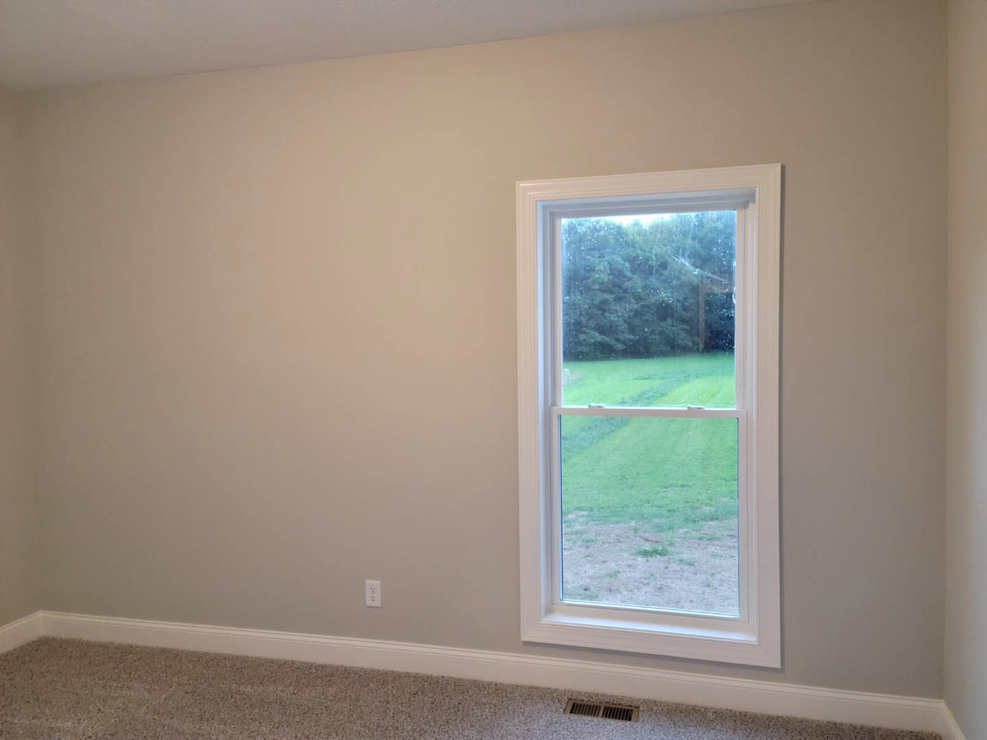 Large window with raindrops overlooking grassy field, white plaster walls, carpeted floor, and white ceiling in residential room