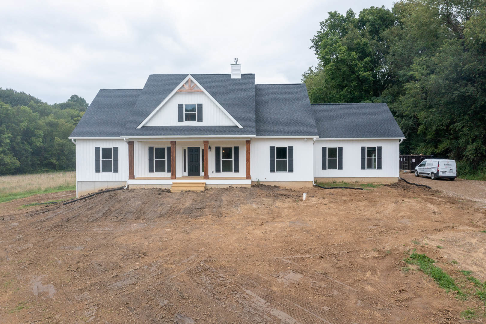 Partially built house with gray roof, black door with white frame, dirt patch in front, surrounding trees, white van parked on nearby road, illuminated window visible