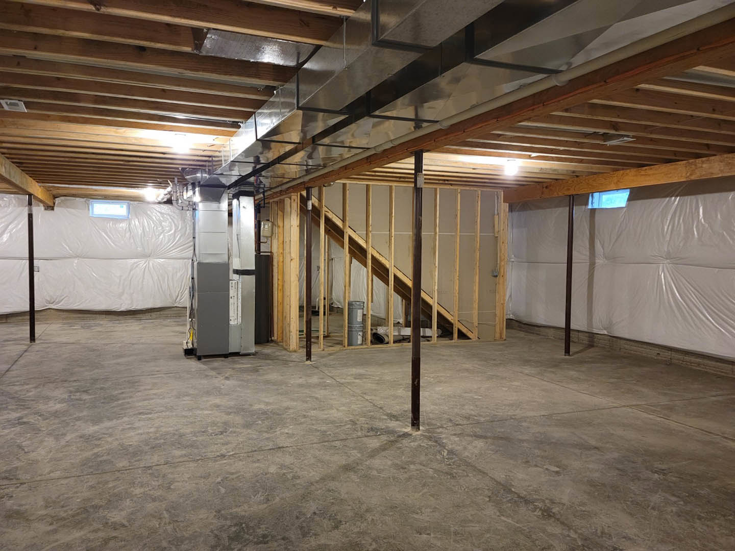 Concrete basement floor with exposed wooden framing, metal support pole, metal staircase, white plastic wall covering, white door with black tape, and metal vent