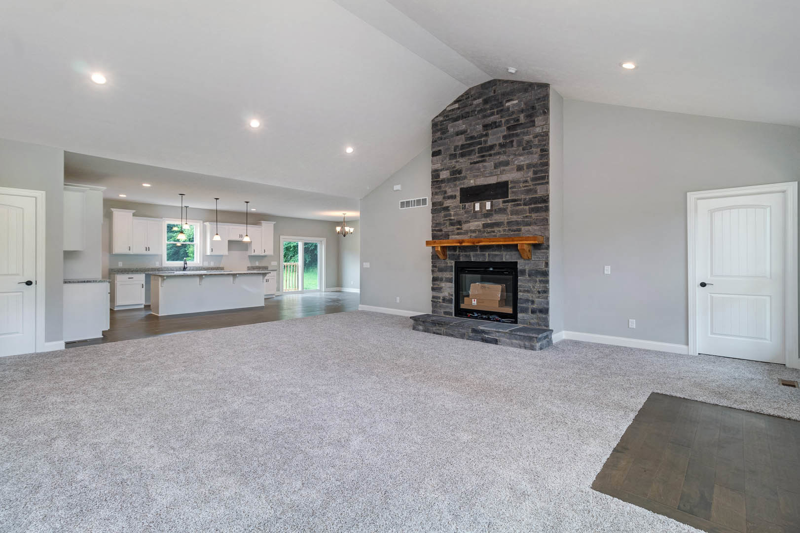 Spacious carpeted living room featuring a fireplace with a wood mantel, adjacent open kitchen with granite countertops, white cabinetry, and a white door with black handle.