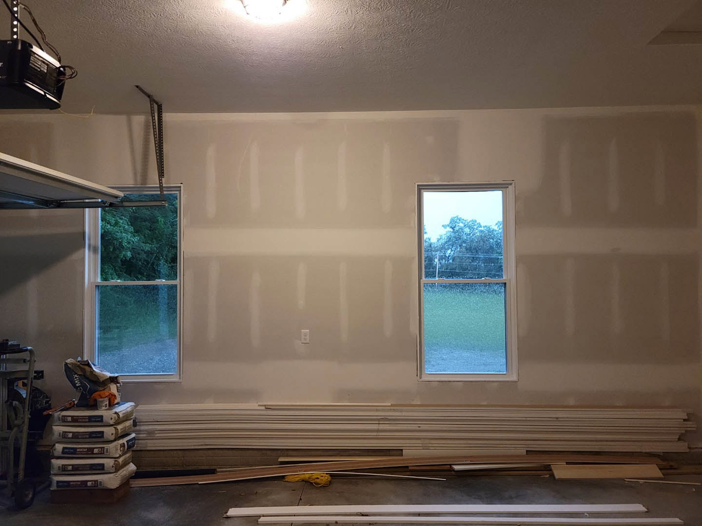 Spacious room with two large windows overlooking grass and trees, unfinished plaster walls, cement bags stacked near the wall, light-colored floor, and a black electrical box