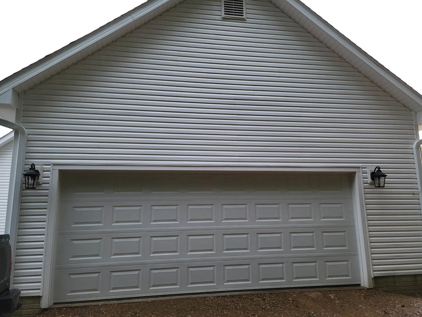 White paneled garage door beneath a gray shingled roof, black lantern mounted beside door, white siding exterior, dirt driveway in foreground