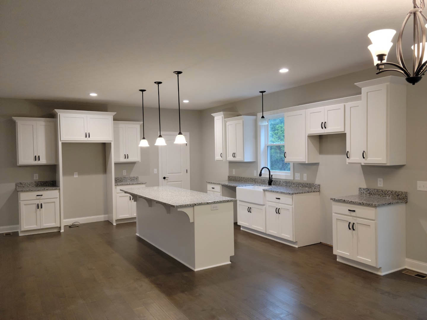 White kitchen with granite countertops, white cabinets featuring black handles, central island, stainless steel sink, wooden flooring with white baseboards, and modern pendant