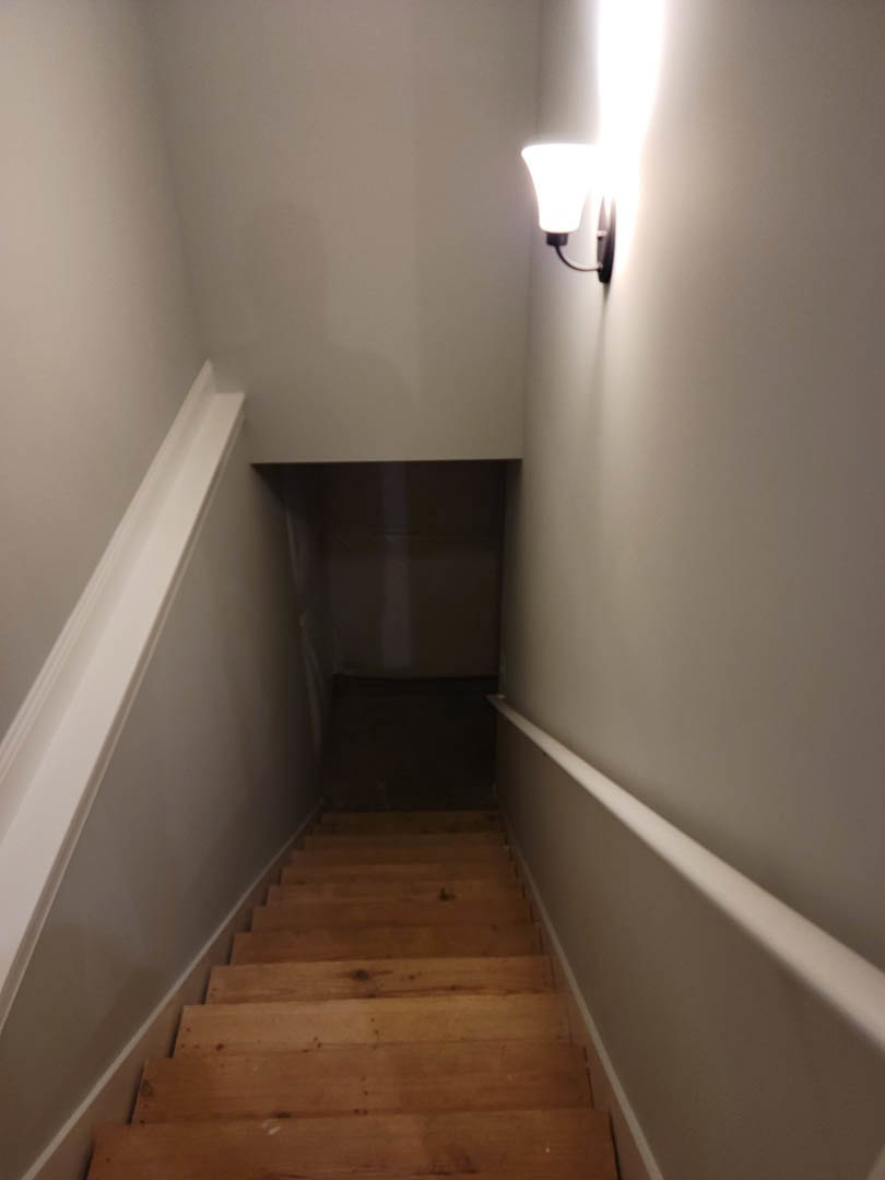 Wooden staircase with integrated wall sconce, white plaster walls, and dark hallway flooring