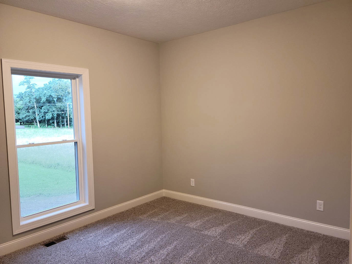 Bedroom with beige carpet, white walls, large window overlooking green field and forest, ceiling vent visible