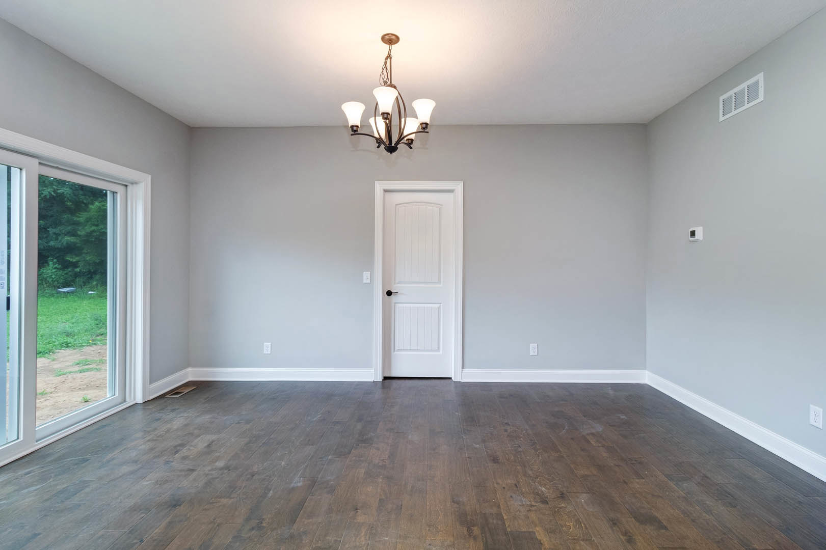 White paneled door with black handle, wood flooring with white baseboard trim, glass door partially open, decorative chandelier hanging from ceiling, wall vent visible.