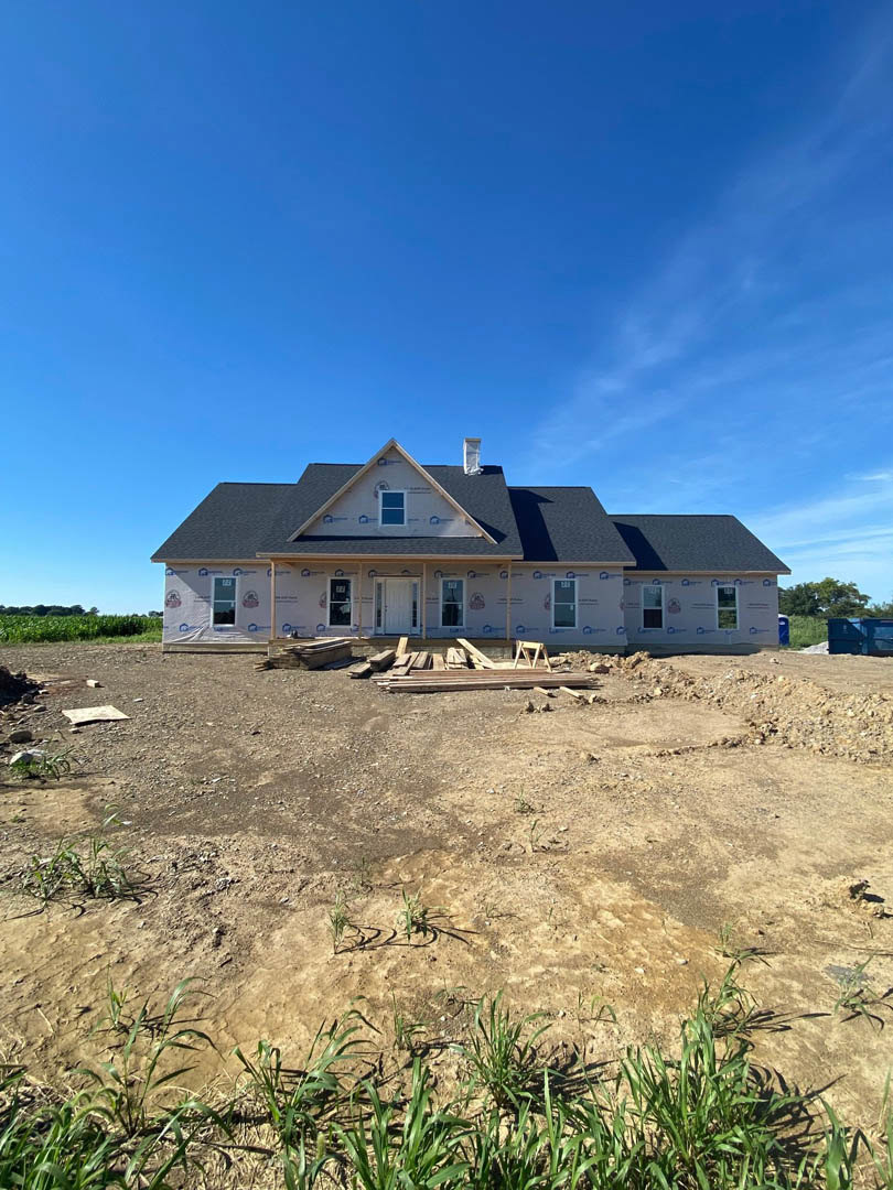 Wood-framed house under construction on dirt lot, white front door installed, blue sky overhead, sparse grass growing around foundation