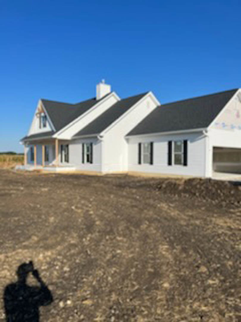 White house with black roof and chimney, partially constructed, surrounded by dirt field; windows installed, siding visible, flag atop roof, clear sky in background.