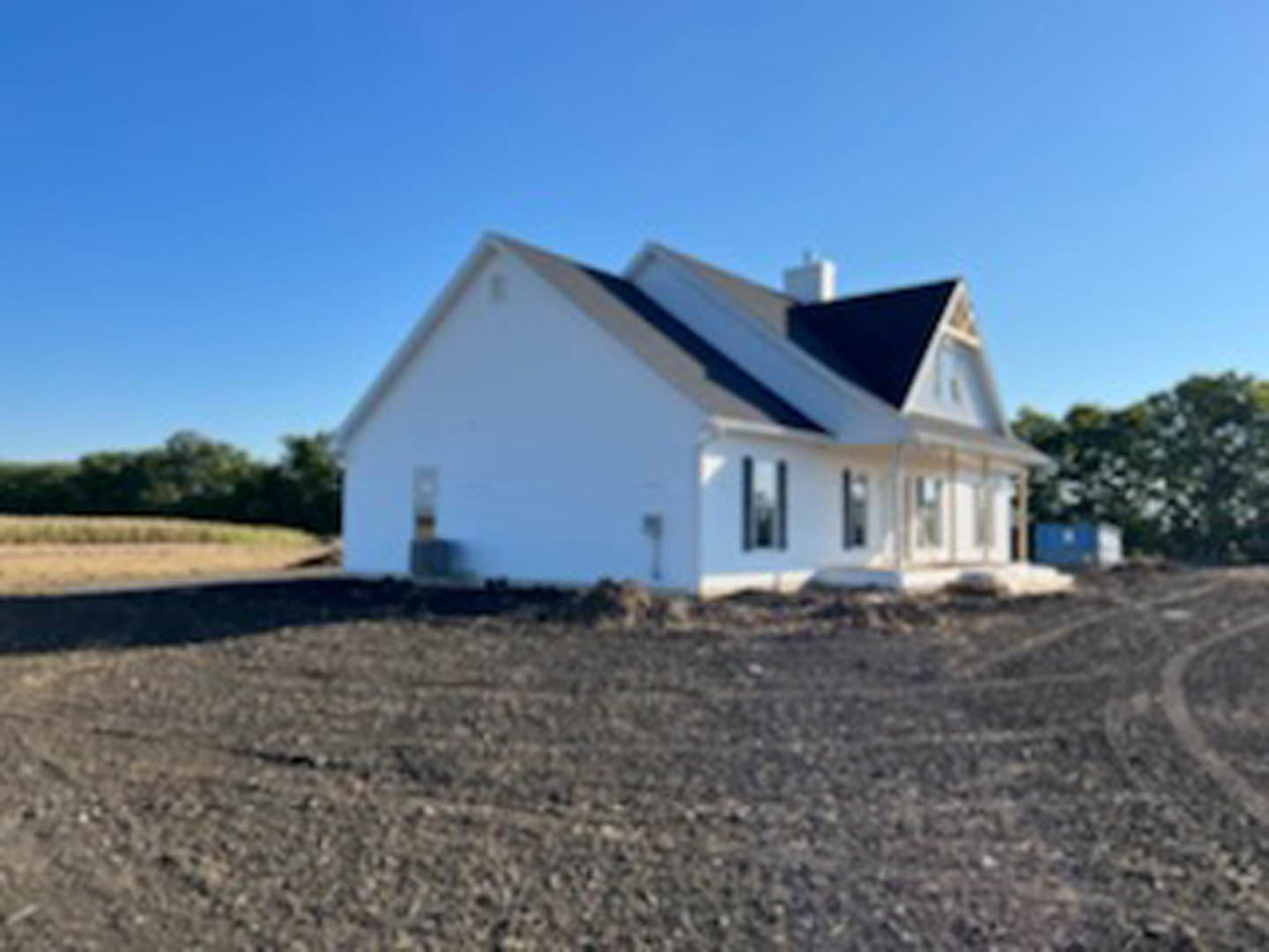 White house with black roof set behind a dirt field, surrounded by open land and sparse trees under a clear sky