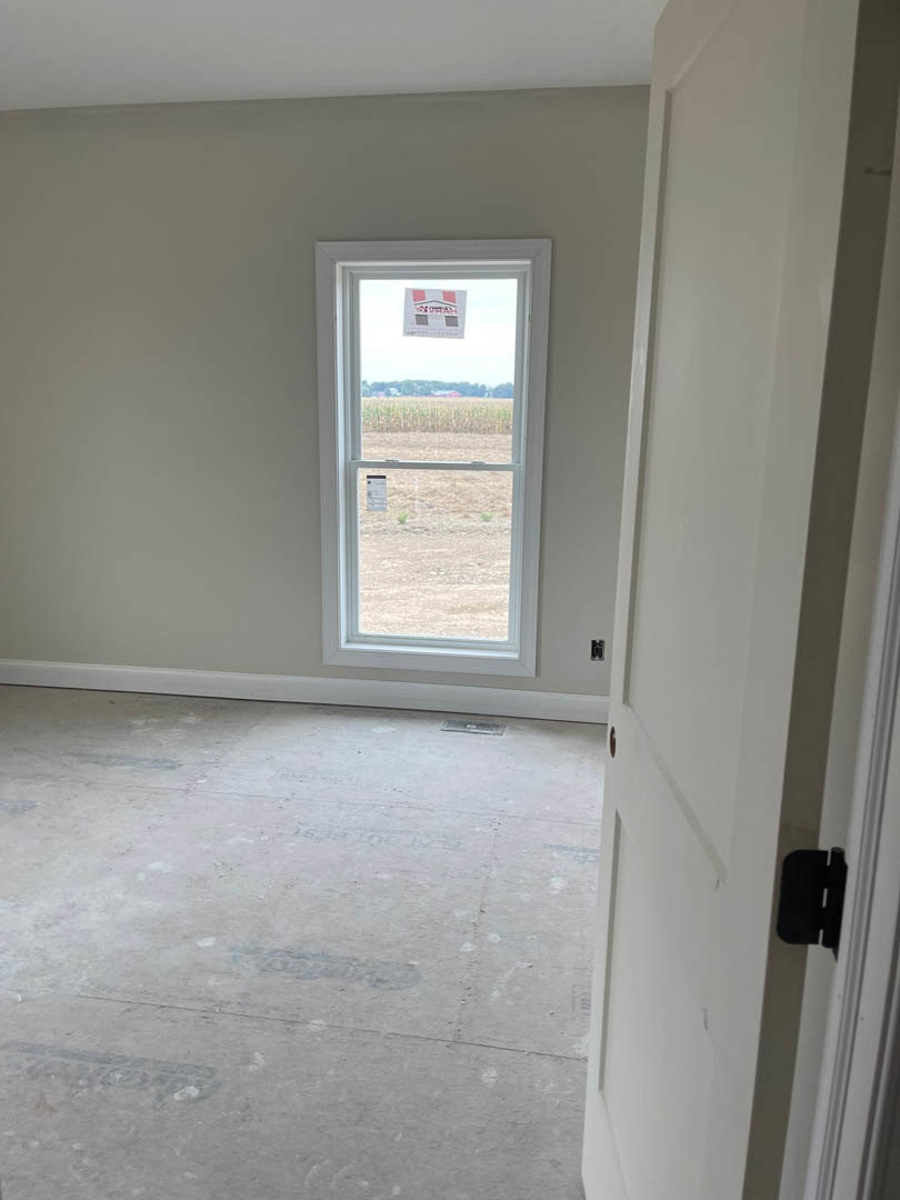 White plaster walls, large window with a black rectangular sign, white door, light wood flooring, and smooth ceiling in a residential interior.