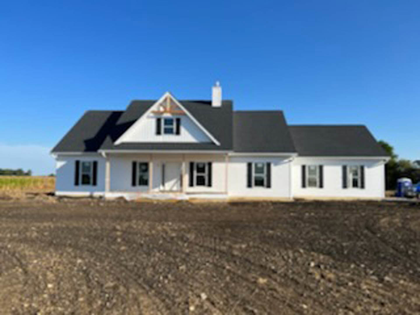 Single-story house with black roof, square windows, and brick chimney, surrounded by dirt field with visible tire tracks under clear sky