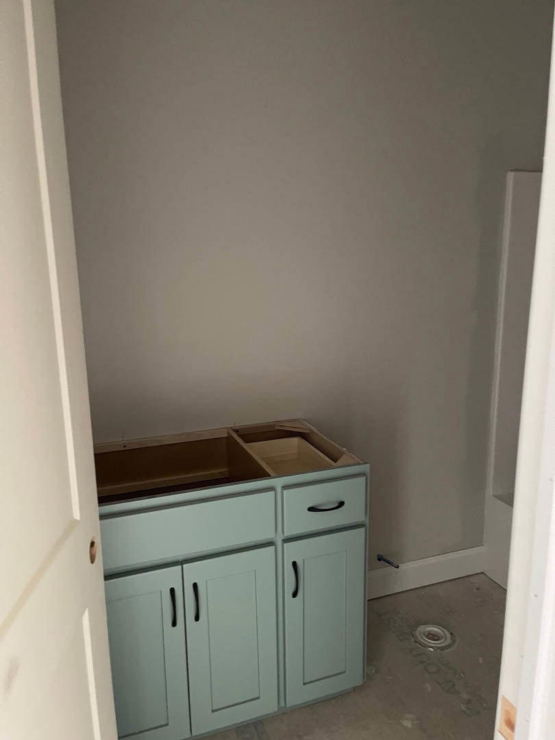 White kitchen cabinet with black handles, multiple drawers, and a circular metal drain visible on the floor; white walls with matching trim.