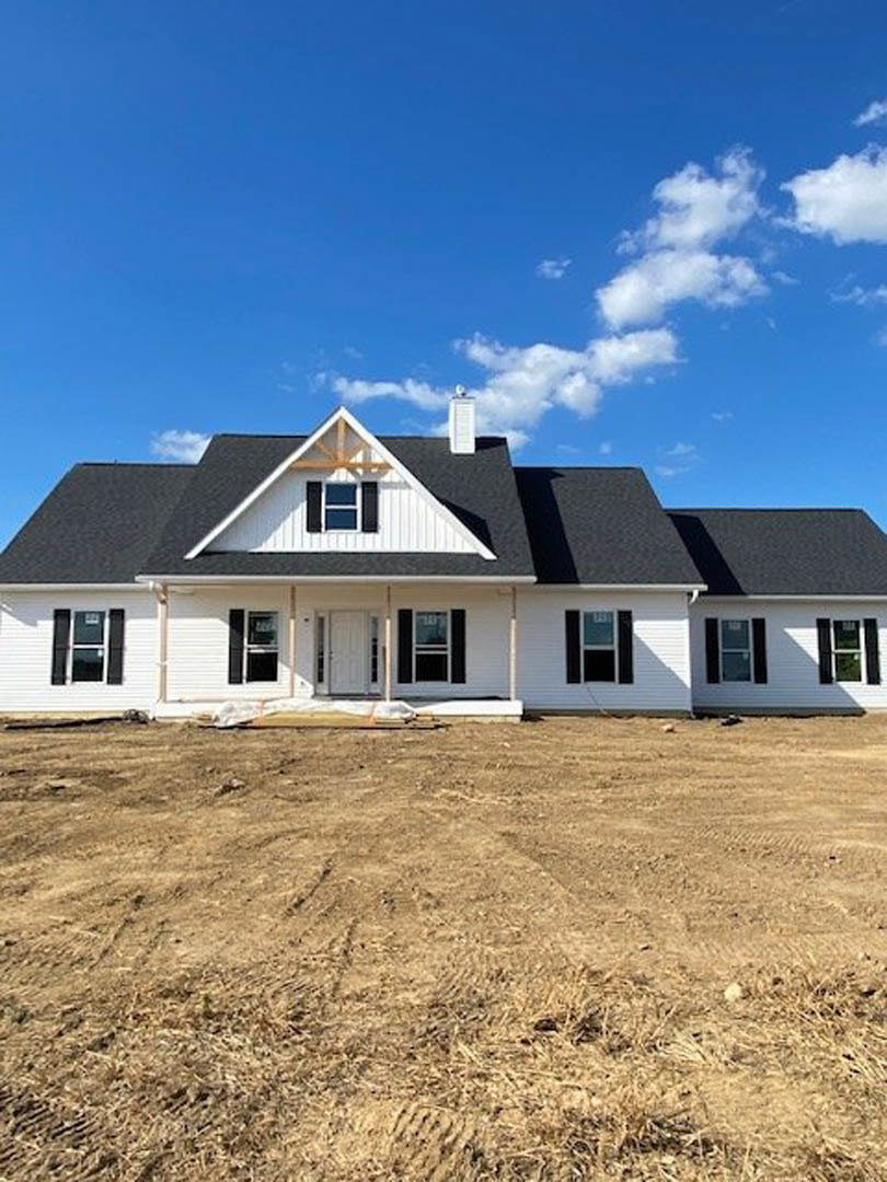 White siding house under construction with black roof, white door, multiple windows, and brown dirt field in foreground